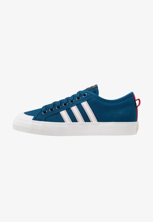 Lage blauwe sneaker met witte rubberen zool, wit ontwerp met drie strepen, donkerblauwe veters en een rood hiel lusje.