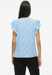 Gaiši zilā blūze ar viļņotiem piedurknēm, sarežģīts ziedu raksts, strukturēta auduma un brīva piegriezuma, skatoties no aizmugures.