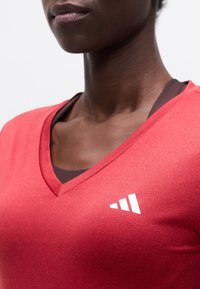 Κοντινό πλάνο ενός ατόμου που φορά ένα κόκκινο μπλουζάκι με V λαιμόκοψη και ένα λευκό τριγωνικό λογότυπο στο στήθος, ντυμένο πάνω από ένα καφέ σκούρο τοπ.