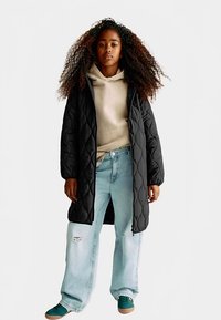 Zwarte doorgestikte lange jas, beige hoodie, lichtblauwe distressed jeans en groene sneakers met een textuur en een wijde pijp. KRULLEND haar zichtbaar.