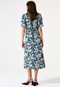 Blumenbedrucktes, türkisfarbenes Midi-Kleid mit kurzen Ärmeln, tailliertem Bund und fließendem Rock. Es werden schwarze Sandalen mit Blockabsatz getragen.