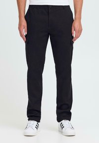 Mann som har på seg svarte rette cargobukser med klafflommer, hvite sneakers med svarte striper, står mot en ensfarget bakgrunn.