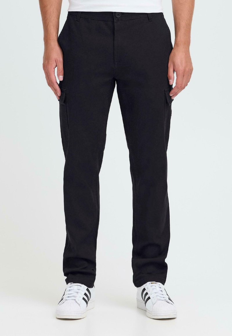 Mann som har på seg svarte rette cargobukser med klafflommer, hvite sneakers med svarte striper, står mot en ensfarget bakgrunn.