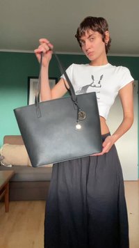 Mala tote em pele sintética preta com duas alças, formato retangular e um detalhamento em forma de porta-chaves. Textura suave, design minimalista.