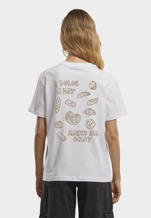 Osoba o długich blond włosach, ubrana w białą koszulkę z nadrukiem szkiców ciast i napisem "A Dolce A Day Makes All Okay" na plecach.