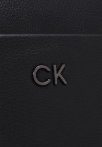 Czarna, teksturowana skóra z metalowym logo "CK" umieszczonym na środku, sugerująca detal modowego akcesorium lub torby.