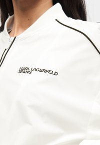 Κοντινό πλάνο λευκού μπουφάν με φερμουάρ και μαύρη επένδυση, με το λογότυπο "KARL LAGERFELD JEANS" στο αριστερό στήθος, φορεμένο από άτομο με μακριά σκουρόχρωμα μαλλιά.