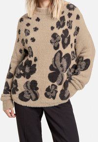Pull beige texturé avec des motifs floraux et abstraits noirs audacieux ; coupe ample avec des poignets retroussés et un col rond.