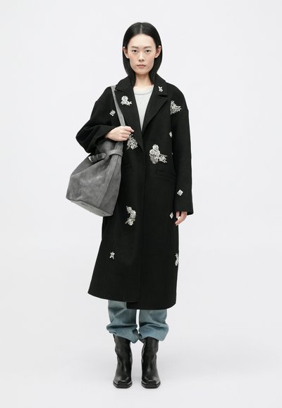 Manteau en laine noir avec broderies florales, design à un bouton, larges revers, poches latérales, associé à un sac en daim gris et un jean en denim.