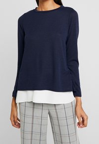 Marinblå långärmad topp i lätt material med rund halsringning. Har sidoklyftor och ett lager på lager-utseende över en vit skjorta.