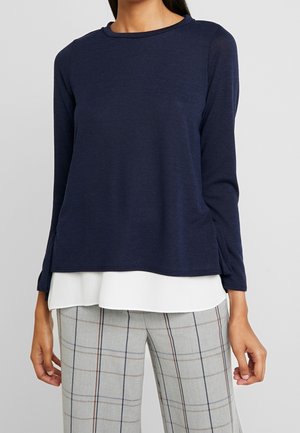 Top de manga larga en color navy, hecho de una tela ligera, con cuello redondo. Cuenta con aberturas en los lados y un diseño de capas encima de una camisa blanca.