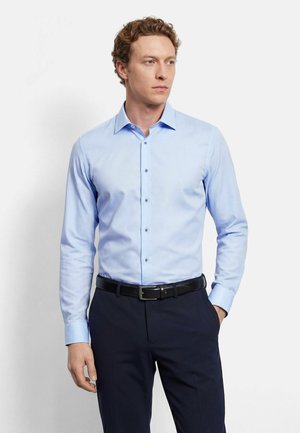 Mann mit lockigem Haar, der ein hellblaues Hemd mit Knöpfen, schwarzen Gürtel und dunkle Hose trägt, steht mit einer Hand in der Tasche.