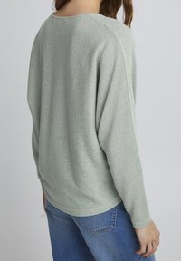 Hellgrüner, strukturierter Strickpullover mit lässiger Passform, fallenden Schultern und langen Ärmeln, kombiniert mit blauen Jeans.