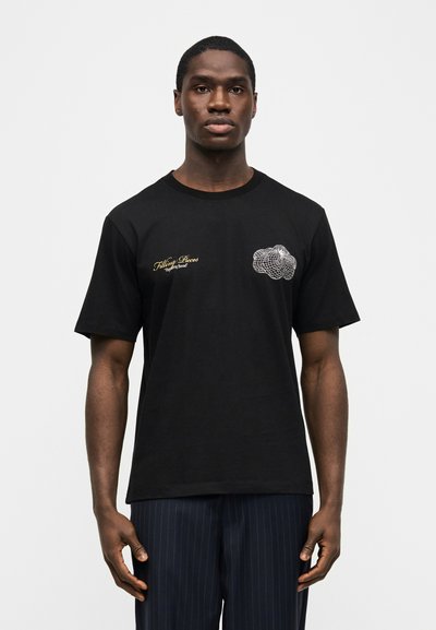 Νεαρός άνδρας με μαύρο t-shirt με μικρό κίτρινο κείμενο και λευκό αφηρημένο σχήμα στο στήθος, που στέκεται μπροστά σε απλό λευκό φόντο.