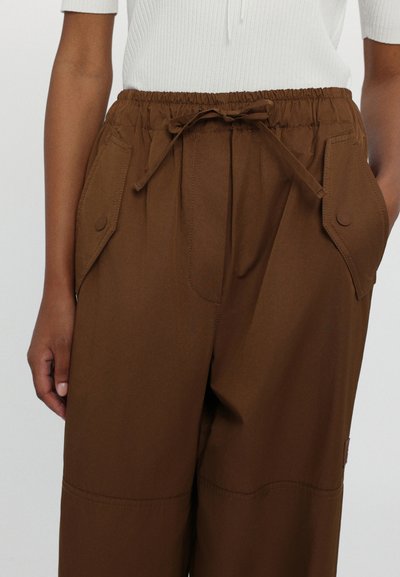 Brown elastic-waist pants feature a drawstring closure, side pockets with flaps, and a smooth texture. Suitable for casual wear.