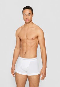 Weiße Baumwoll-Boxershorts mit einem dehnbaren Bund, der ein subtileres Logo-Detail aufweist. Modell steht vor einem neutralen grauen Hintergrund.