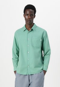 Camisa azul claro de botones con un bolsillo en el pecho izquierdo, mangas largas y una textura suave, llevada sobre pantalones grises.