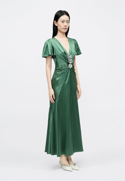 Rochie verde din satin, cu mâneci scurte, evazate, decolteu adânc și ornament central. Lungime până în podea, cu o siluetă fluidă.