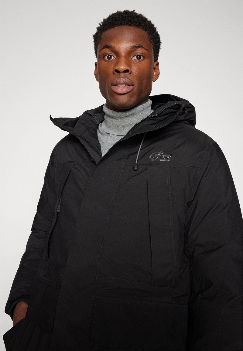 Hombre con chaqueta de invierno con capucha negra sobre cuello de tortuga gris, mirando hacia adelante contra un fondo simple.