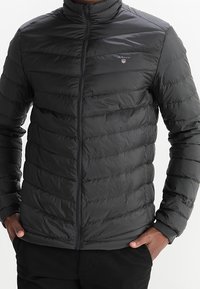 Tumma harmaa untuvatakki, jossa on pystysuorat tikatut osat, korkea kaulus, täysi etuvetoketju ja logo vasemmassa rinnassa. Sileä, kevyt materiaali.