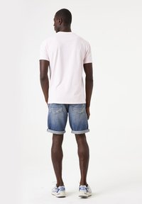 T-shirt en coton rose clair, shorts en jean bleu avec des poignets roulés et chaussures de sport grises avec des accents bleus. Le modèle est vu de dos.