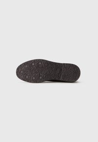 Semelle de chaussure noire texturée avec des motifs de grip circulaires et logo de la marque visible sur un fond clair.