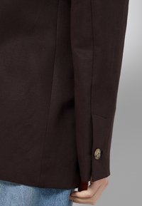 Manche de blazer marron avec un poignet boutonné portée sur un jean en denim bleu clair, avec une main détendue sur le côté sur un fond neutre.