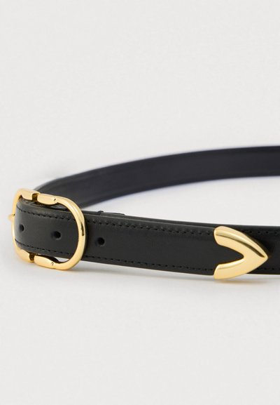 Μαύρη δερμάτινη ζώνη με λεία υφή, που διαθέτει πόρπη σε χρυσό τόνο και μεταλλικό διακοσμητικό σχήματος V. Ρυθμιζόμενη με πολλαπλές τρύπες.