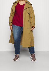 Manteau beige avec des boutons et une ceinture, associé à un pull rouge et un jean bleu. Le modèle porte des baskets marron et blanches.