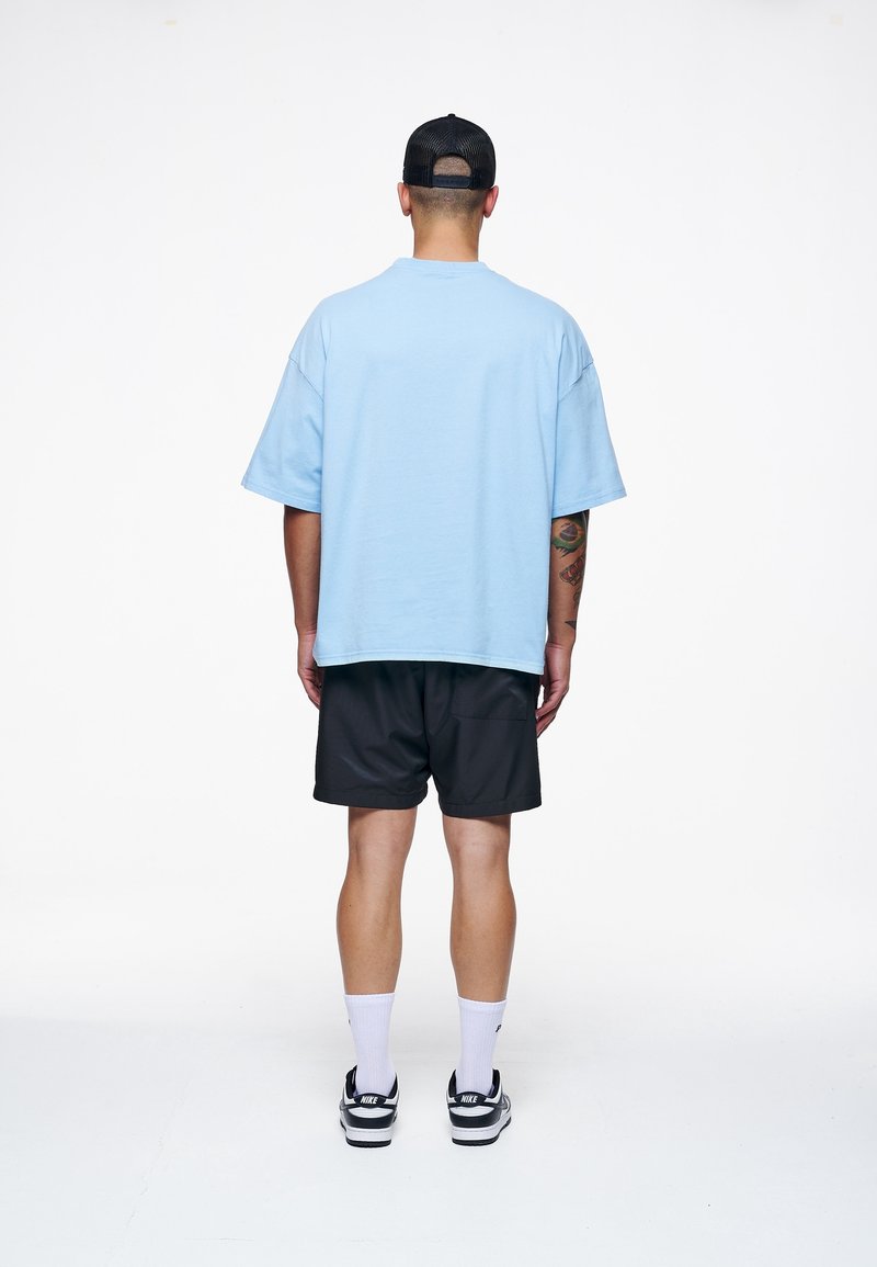 Homme portant un t-shirt bleu clair oversize, un short noir, des chaussettes blanches, des baskets Nike noires et une casquette noire, debout de dos sur fond blanc.