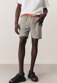 Pantalones cortos de toalla gris con un cordón rosa y acentos en verde neón, combinados con sandalias negras. El modelo está de pie sobre un suelo gris claro.