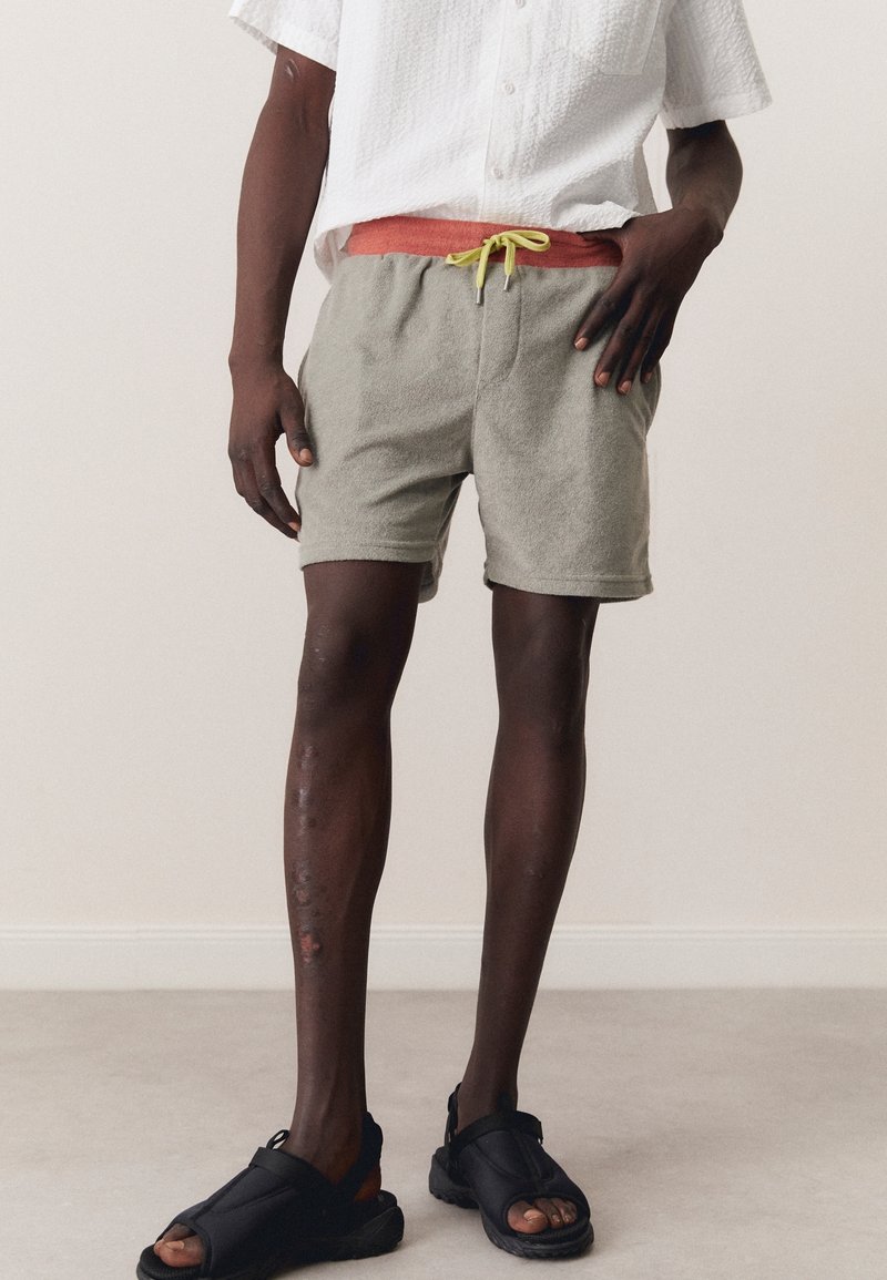 Pantalones cortos de toalla gris con un cordón rosa y acentos en verde neón, combinados con sandalias negras. El modelo está de pie sobre un suelo gris claro.