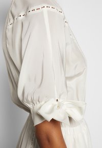 Manche de blouse blanche avec détail bouffant et nœud décoratif noué au poignet, présentant une couture ajourée près de l'épaule.