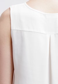 Bílá bezrukávová blůza s hladkou texturou, kulatým výstřihem a jednoduchým skladem na zadní straně pro zvýraznění designu.