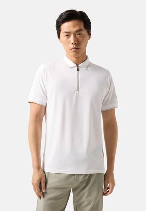 Ung mand iført en hvid kortærmet polo med lynlås og lyse beige bukser mod en ensfarvet baggrund.