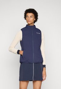 Colete sem mangas azul-marinho com gola alta, fecho éclair frontal e logo da Calvin Klein. Textura suave, design ajustado, combinado com uma blusa de manga longa leve.