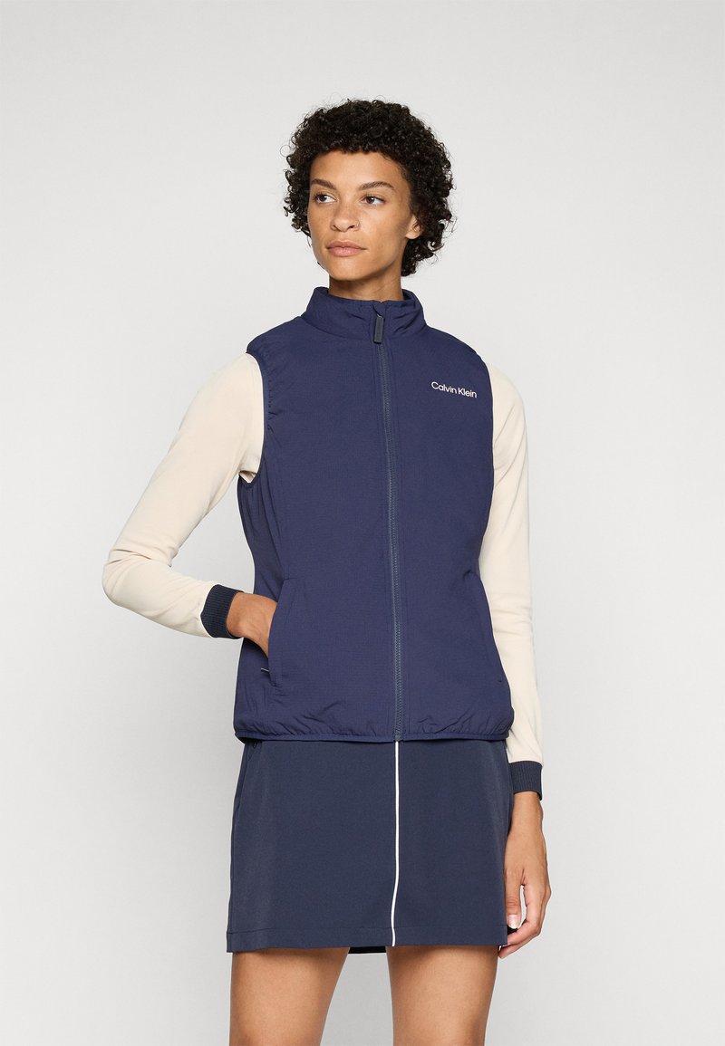 Colete sem mangas azul-marinho com gola alta, fecho éclair frontal e logo da Calvin Klein. Textura suave, design ajustado, combinado com uma blusa de manga longa leve.