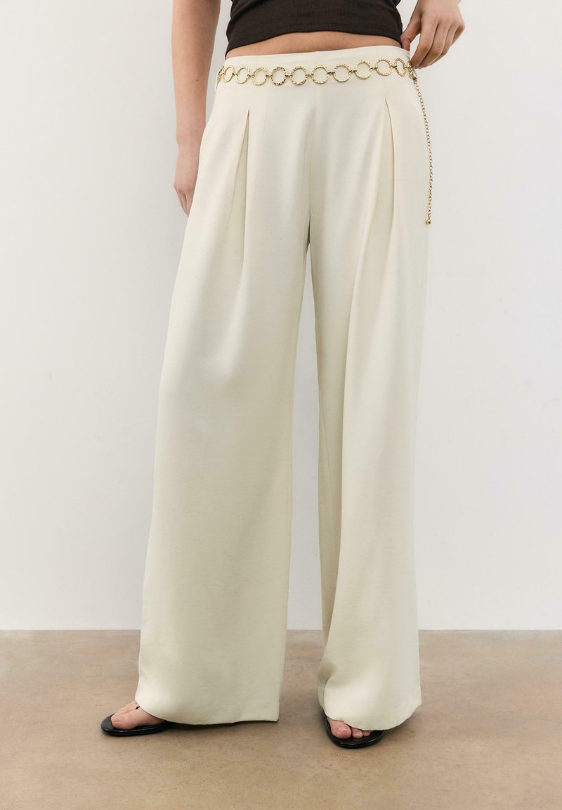 Femme portant un pantalon large crème, un crop top noir, des sandales noires et une ceinture chaîne dorée à maillons circulaires, debout devant un fond uni.