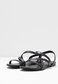 Sandales plates noires avec plusieurs fines lanières en cuir croisées sur le pied et une lanière à l'arrière, posées sur une surface blanche.