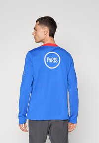 Plava majica dugih rukava s crvenim ovratnikom, sa bijelim kružnim logotipom na leđima koji sadrži natpis "PARIS". Glatka tkanina, atletski kroj, minimalistički dizajn.