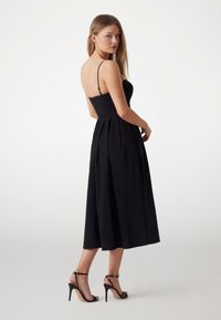 Robe noire midi avec bretelles fines, corsage ajusté et jupe plissée. Portée avec des sandales à talons hauts noires. Texture de tissu lisse.