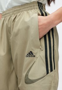 Bézs színű sportnadrág elasztikus derékrésszel, fekete Adidas logóval és három fekete függőleges csíkkal az oldalakon. Simogató textúra, könnyű anyag.
