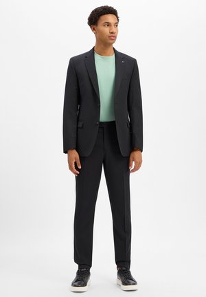 Young man standing wearing a black suit with a mint green t-shirt and black sneakers against a plain white background.