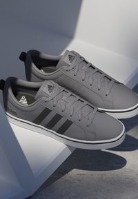 Zapatillas bajas grises de Adidas con rayas negras y suelas blancas, exhibidas sobre superficies blancas inclinadas contra un fondo gris.