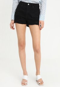 Pantalones cortos de mezclilla negra con dobladillo deshilachado y detalles rasgados, combinados con una camisa de manga larga de lunares azul claro y sandalias blancas de dedo.