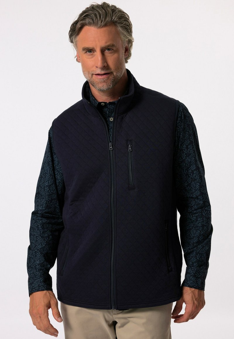 Mörkblå quiltad väst med hög krage, med blixtlåsfickor och en mönstrad grön skjorta under. Slätt tyg, skräddarsydd passform.