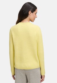 Żółty sweter prążkowany z długimi rękawami i okrągłym dekoltem, o miękkiej fakturze. Noszony z w kratkę spodniami, pokazujący widok z tyłu.