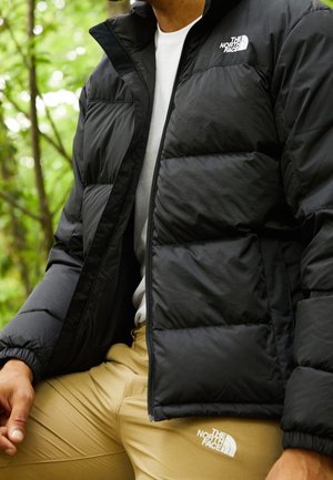 Mężczyzna ubrany w czarną kurtkę puchową i beżowe spodnie z logo The North Face, siedzący na zewnątrz na tle zielonych liści.