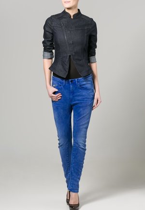 Chaqueta ajustada de denim oscuro con cuello mao, cierre de botones y mangas enrolladas en contraste; combinada con jeans ajustados azules y tacones.
