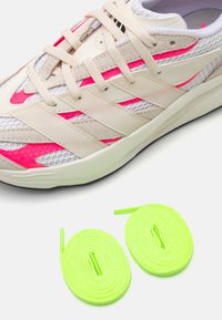 Zapatilla deportiva beige con acentos de malla y gamuza, que presenta rayas rosas y cordones crema. Incluye dos cordones de color verde neón cercanos.